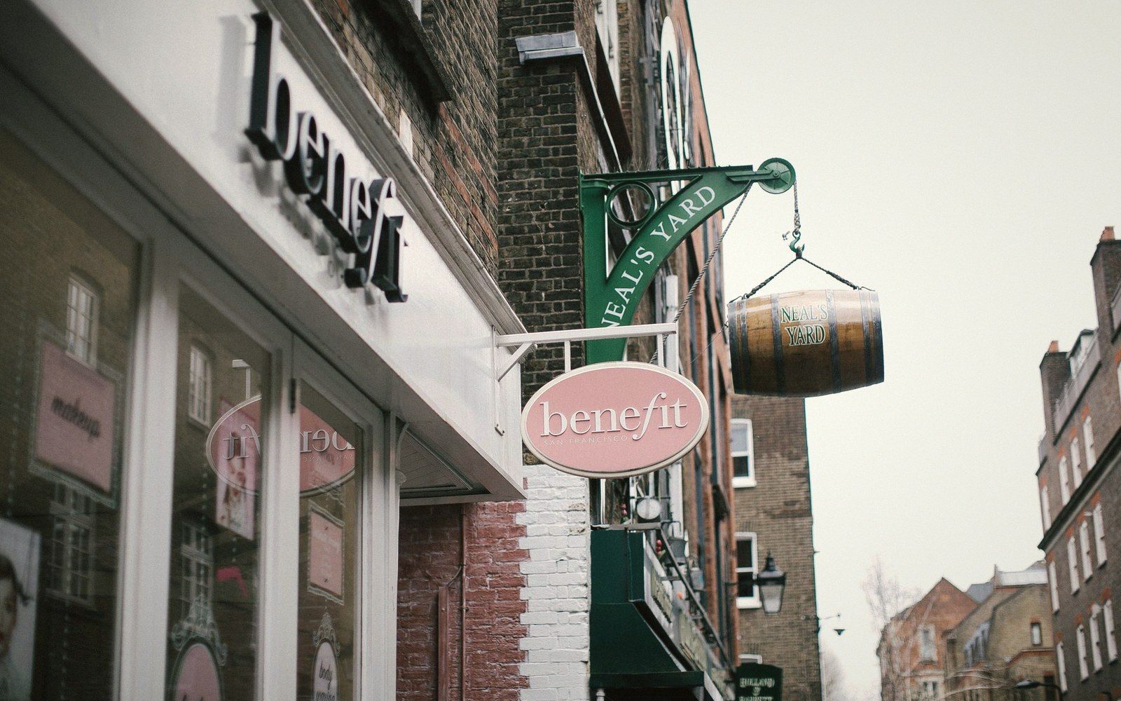 Street-level view of a row of Indian-owned shops on a British high street, hand-painted signage in English and Gujarati above the awnings